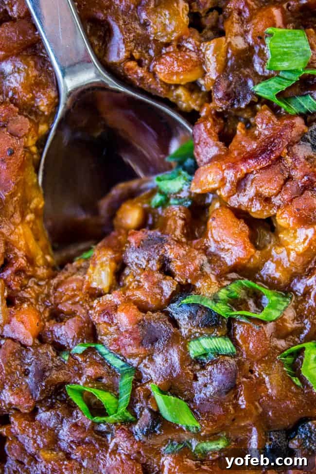 Crispy Bacon Bean Bake 3 A serving spoon in a rustic pan filled with bubbling, hearty bacon and beef baked beans, ready to be served.