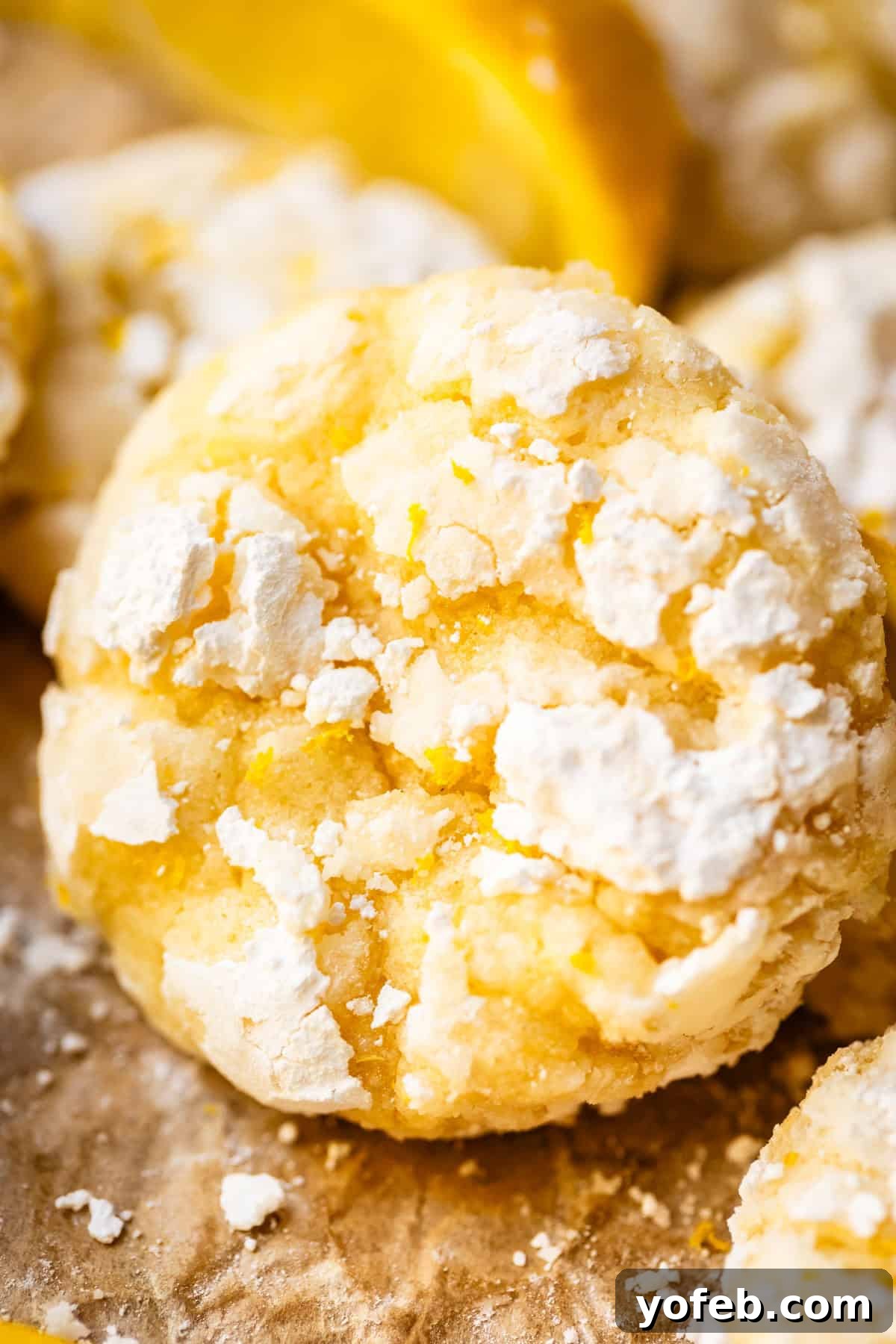 Zesty Lemon Crinkles 2 Close-up of a lemon crinkle cookie covered in powdered sugar, with a cracked surface and a bright yellow color. Blurred lemon slices and more cookies are visible in the background, hinting at fresh ingredients and more delicious treats.