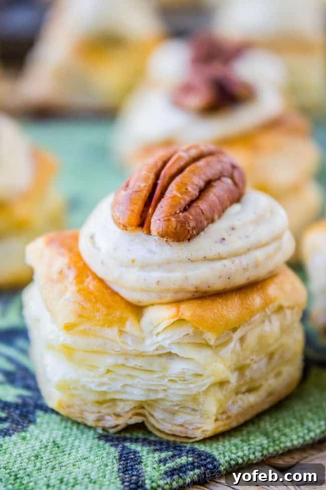 Pillowy Pumpkin Cream Cheese Puffs 2 A close-up of a puff pastry bite topped with creamy pumpkin frosting and a pecan half, set on a green patterned cloth with more pastries blurred in the background.