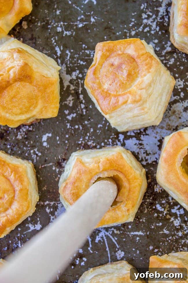 Pillowy Pumpkin Cream Cheese Puffs 8 Hexagon-shaped pieces of golden puff pastry with hollow centers are arranged on a dark baking sheet. A wooden handle is inserted into the center of one pastry.