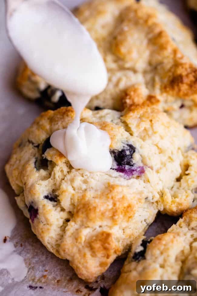A generous drizzle of sweet glaze cascades over a warm blueberry scone from a spoon, promising a delightful finish.