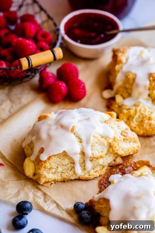 Glazed scones beautifully arranged on parchment paper, adorned with fresh raspberries and a jar of homemade jam in the soft background.