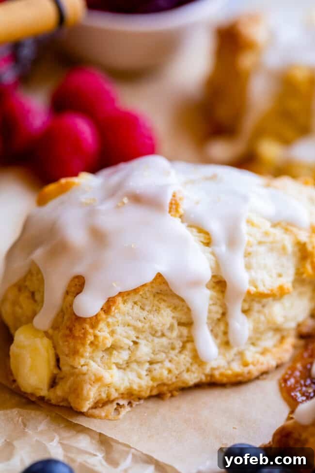 A single glazed scone, glistening invitingly on parchment paper, surrounded by fresh, plump blueberries, highlighting its delicious potential.