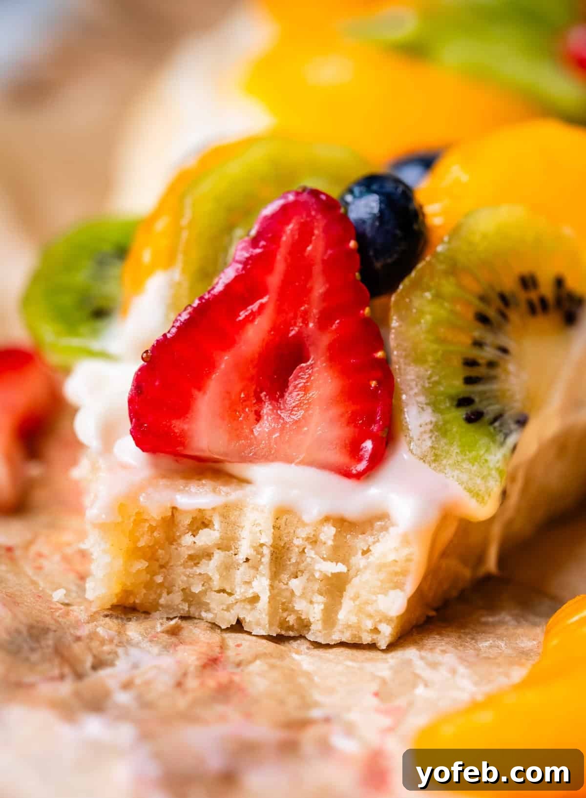 Pillowy Sugar Cookie Fruit Pizza Delight 3 A close-up shot of a slice of fruit pizza with a bite taken, revealing its layers of soft cookie crust, creamy frosting, and colorful fruit toppings.