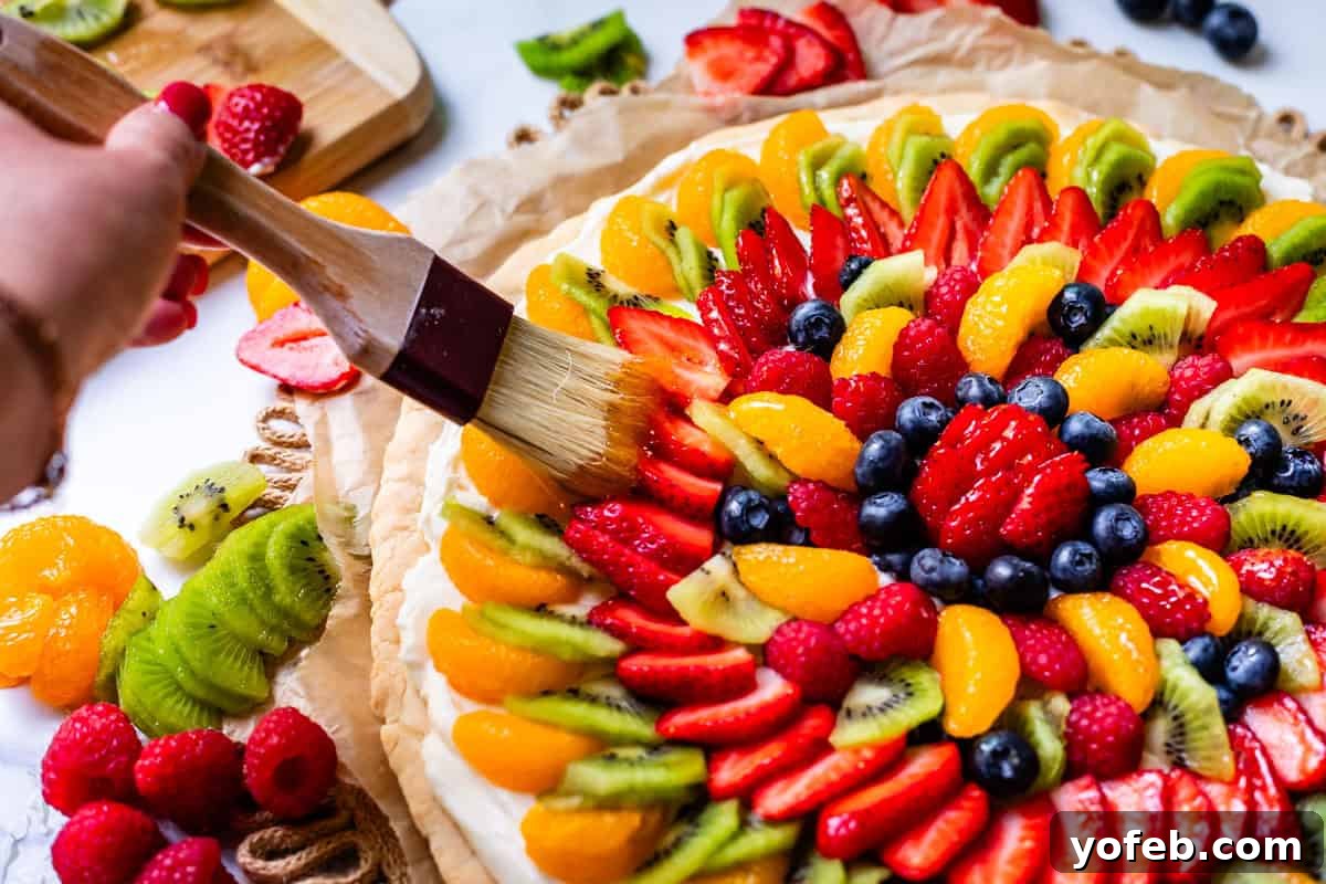 Pillowy Sugar Cookie Fruit Pizza Delight 21 A hand using a pastry brush to lightly glaze the fresh fruit on top of a baked and frosted sugar cookie pizza.