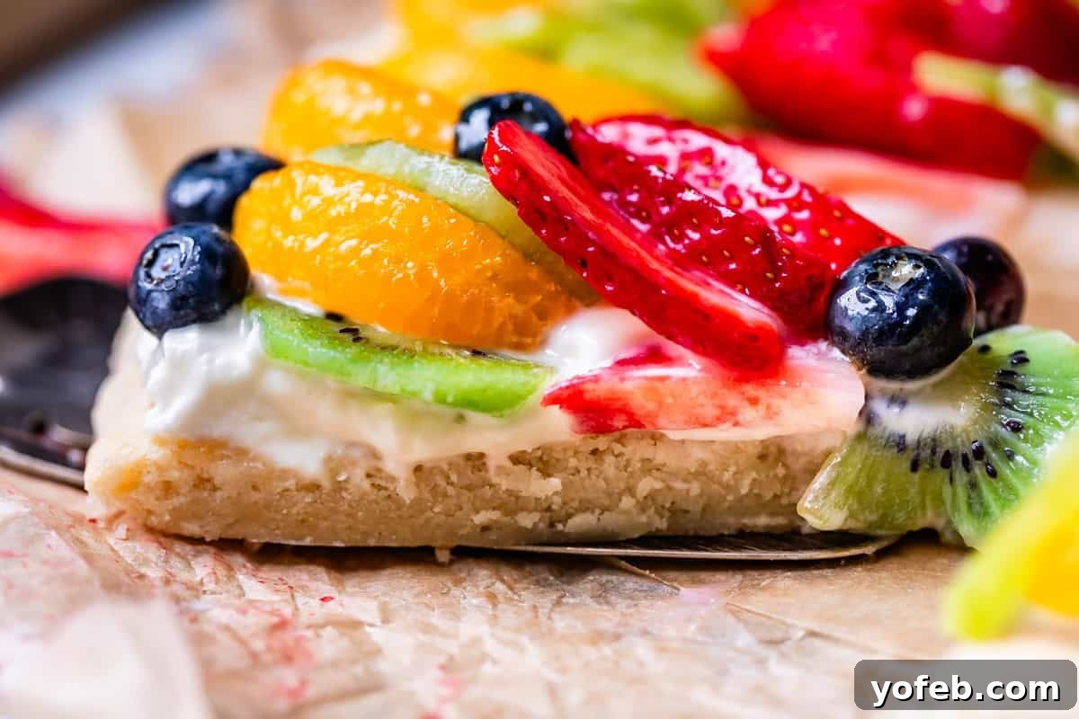 Pillowy Sugar Cookie Fruit Pizza Delight 6 A close-up, side view of a slice of fruit pizza, revealing the distinct layers of the tender sugar cookie base, creamy white frosting, and colorful fresh fruit toppings.