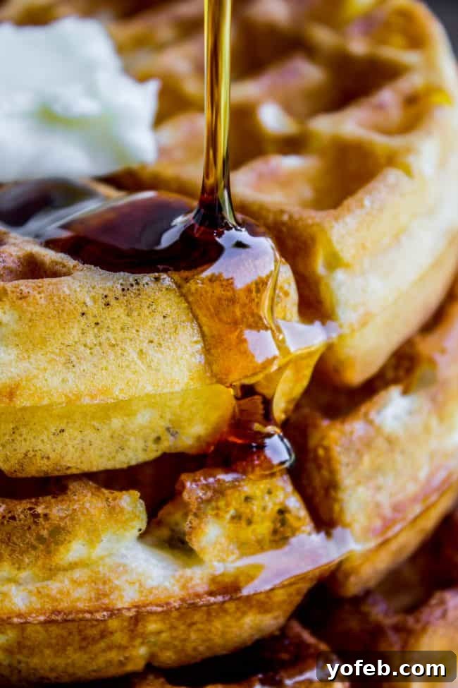 Close-up of syrup being poured over a stack of golden waffles topped with a dollop of whipped butter.