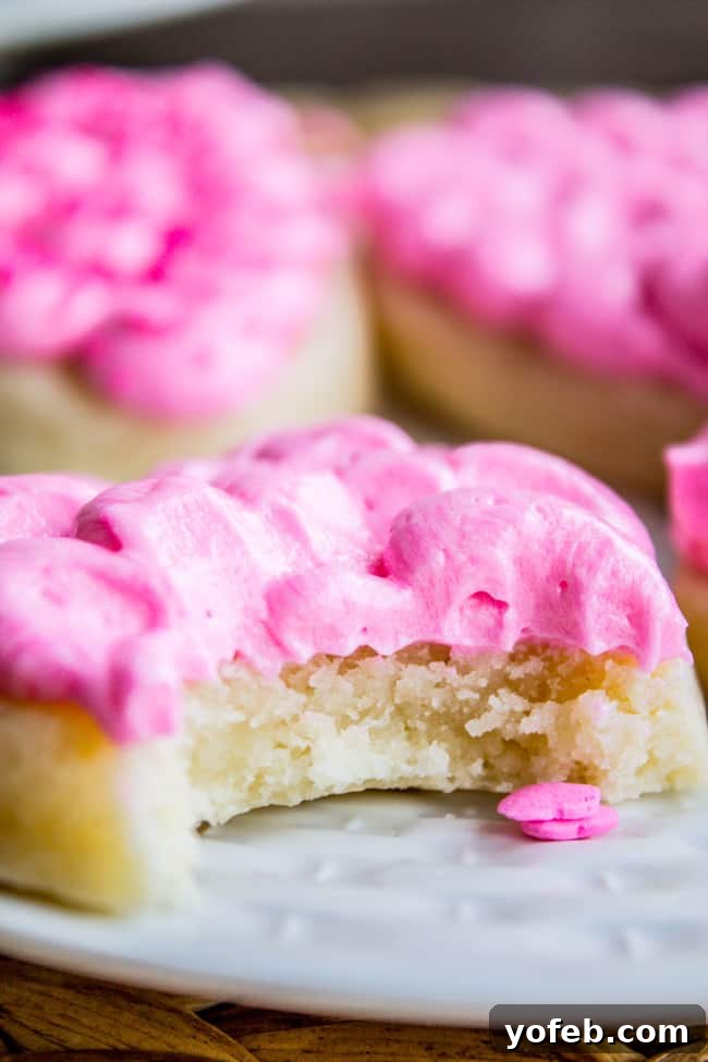 THICK SOFT SUGAR COOKIE RECIPE THAT HOLDS ITS SHAPE 3 A soft sugar cookie with thick, pink frosting sits on a plate, a bite revealing its moist, dense interior. Other similarly frosted cookies are blurred in the background.