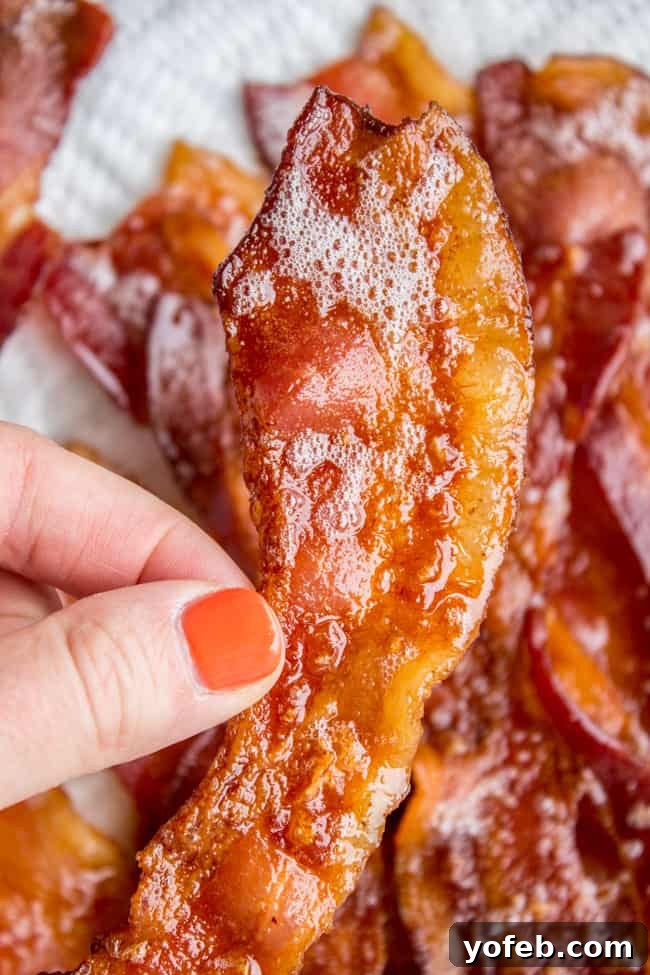 How to Bake Bacon in the Oven in 12 Minutes from the Food Charlatan. I literally never make bacon on the stove anymore. It is SO much easier cooking bacon in the oven than to stand over a hot stove getting splattered with grease. And you can make a whole pan at a time, making it great for a crowd. You can even make it ahead! Let me show you how to bake bacon - it's simple. #bakingbacon #bacon #intheoven #howto #crispy #breakfast #simplerecipe #cookiesheets #350 #lowcarb #comfortfoods A hand holding a piece of perfectly crispy oven-baked bacon, with more strips visible on a sheet pan.