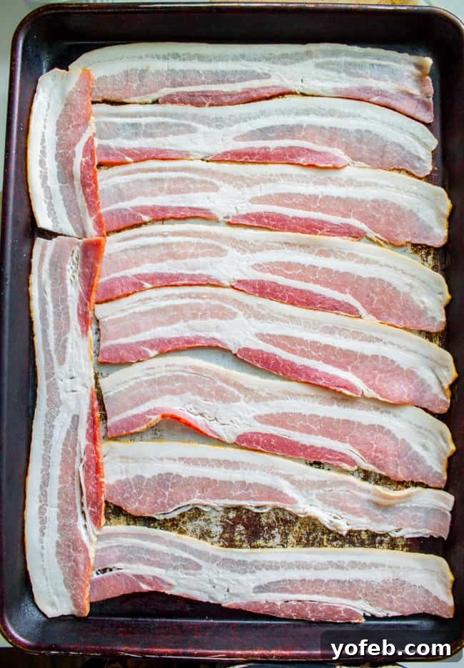 How to Bake Bacon in the Oven in 12 Minutes from the Food Charlatan. I literally never make bacon on the stove anymore. It is SO much easier cooking bacon in the oven than to stand over a hot stove getting splattered with grease. And you can make a whole pan at a time, making it great for a crowd. You can even make it ahead! Let me show you how to bake bacon - it's simple. #bakingbacon #bacon #intheoven #howto #crispy #breakfast #simplerecipe #cookiesheets #350 #lowcarb #comfortfoods Raw bacon strips neatly arranged in a single layer on a baking sheet, ready for the oven.