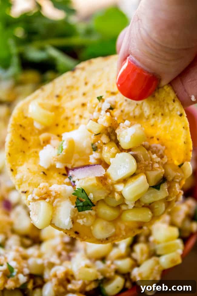 Mexican Street Corn Dip (Elote) from The Food Charlatan. This corn dip recipe is just like an elote (Mexican street corn). It is the perfect combo of sweet corn and savory tart cheese. Plus it's a lot easier to eat AND you get tortilla chips. Win! #corn #dip #elote #mexican #streetfood #summerfood #4thofjuly #cincodemayo #cotija #cornonthecob A hand enthusiastically scooping up Mexican street corn dip with a crunchy tortilla chip.