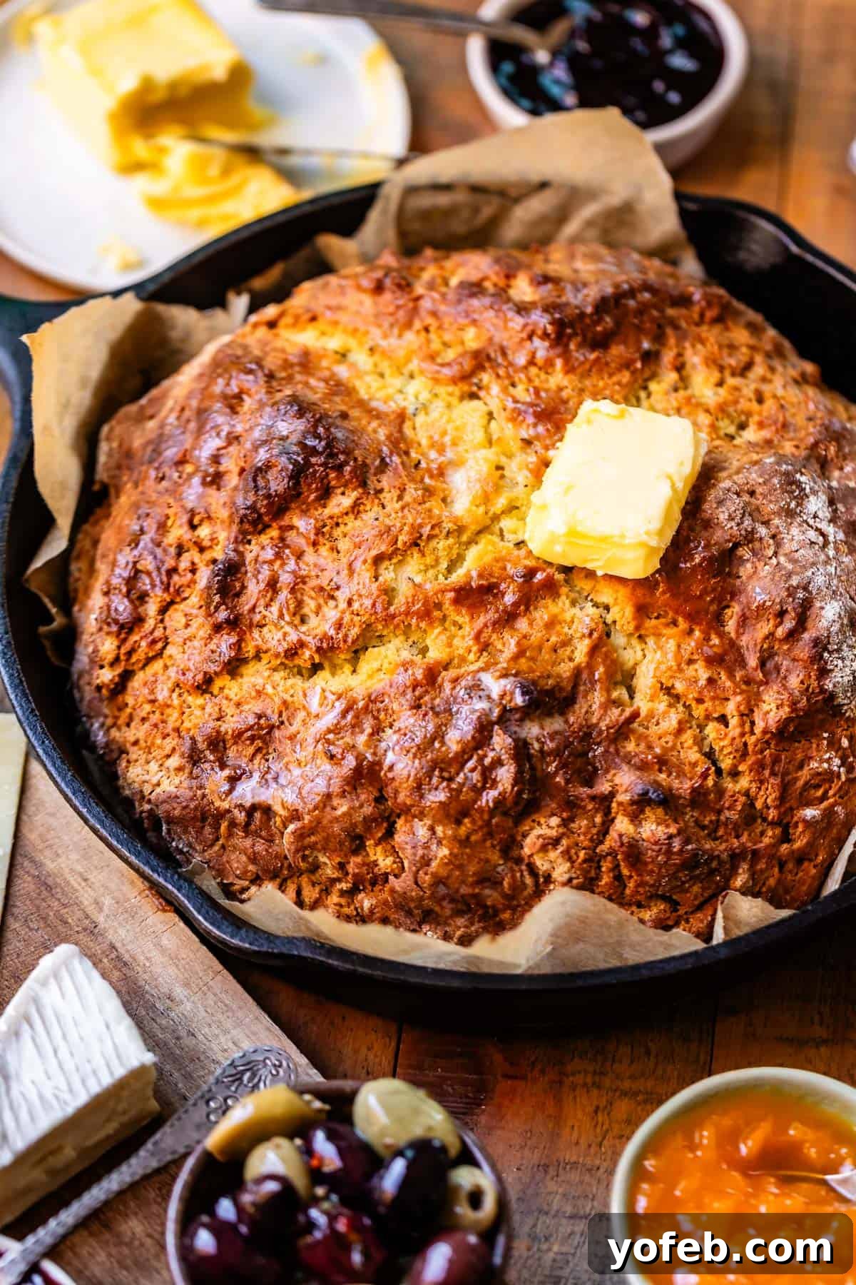 Pure Irish Soda Bread Perfection 3 Baked round loaf of Irish soda bread with golden brown crusty top on parchment paper in cast iron skillet with toppings surrounding including butter, jam, and olives.
