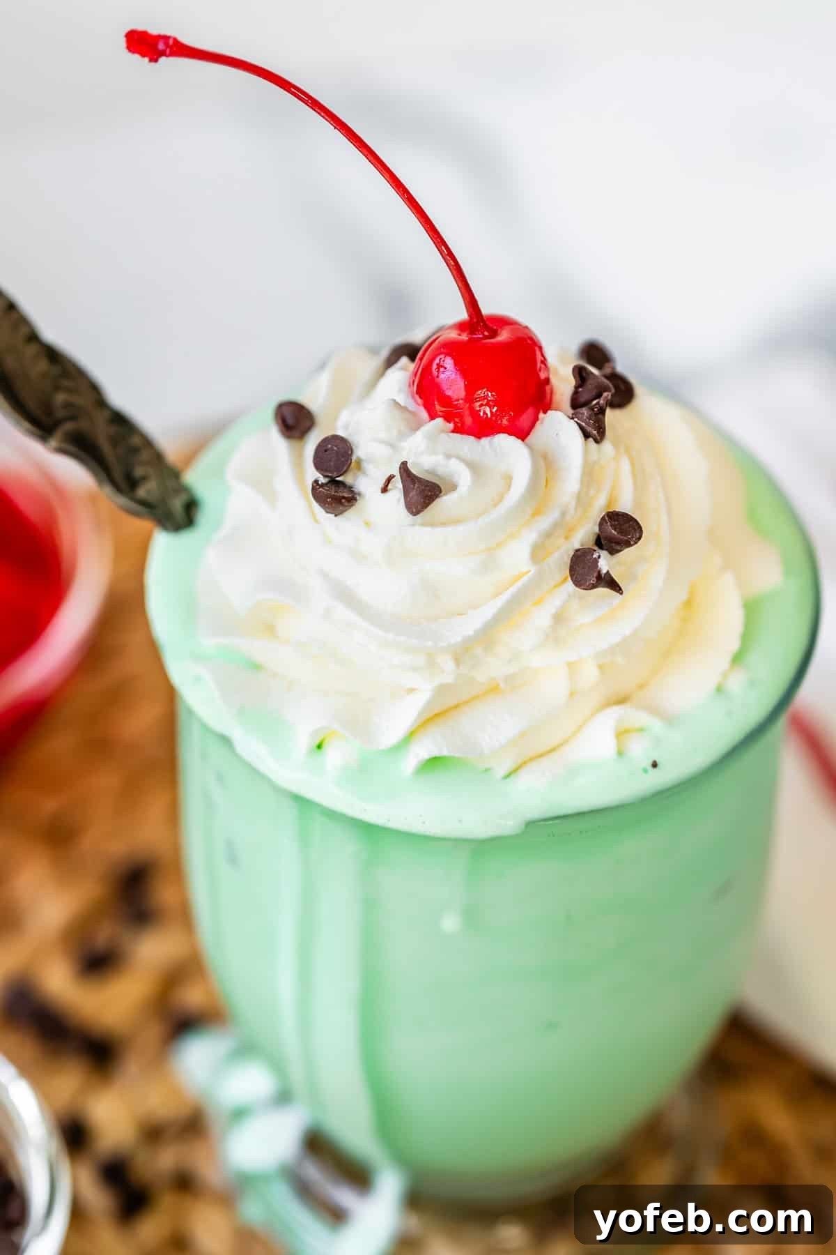 Leprechaun's Delight Shake 17 A glass containing a creamy green shake, lavishly topped with piped whipped cream, a vibrant maraschino cherry, and mini chocolate chips for an extra touch of indulgence.
