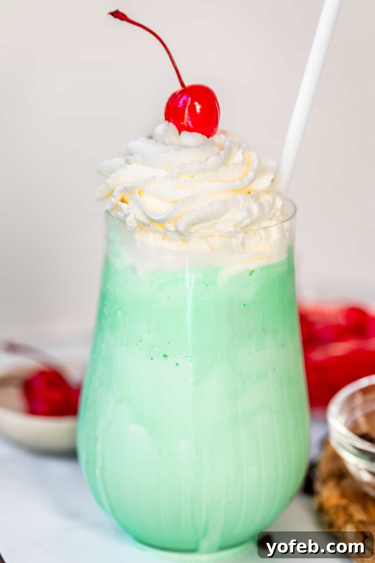 Leprechaun's Delight Shake 3 A tall, fluted glass cup brimming with a vibrant green Shamrock Shake, beautifully topped with a swirl of whipped cream and a bright red maraschino cherry. The shake's texture appears smooth and creamy.