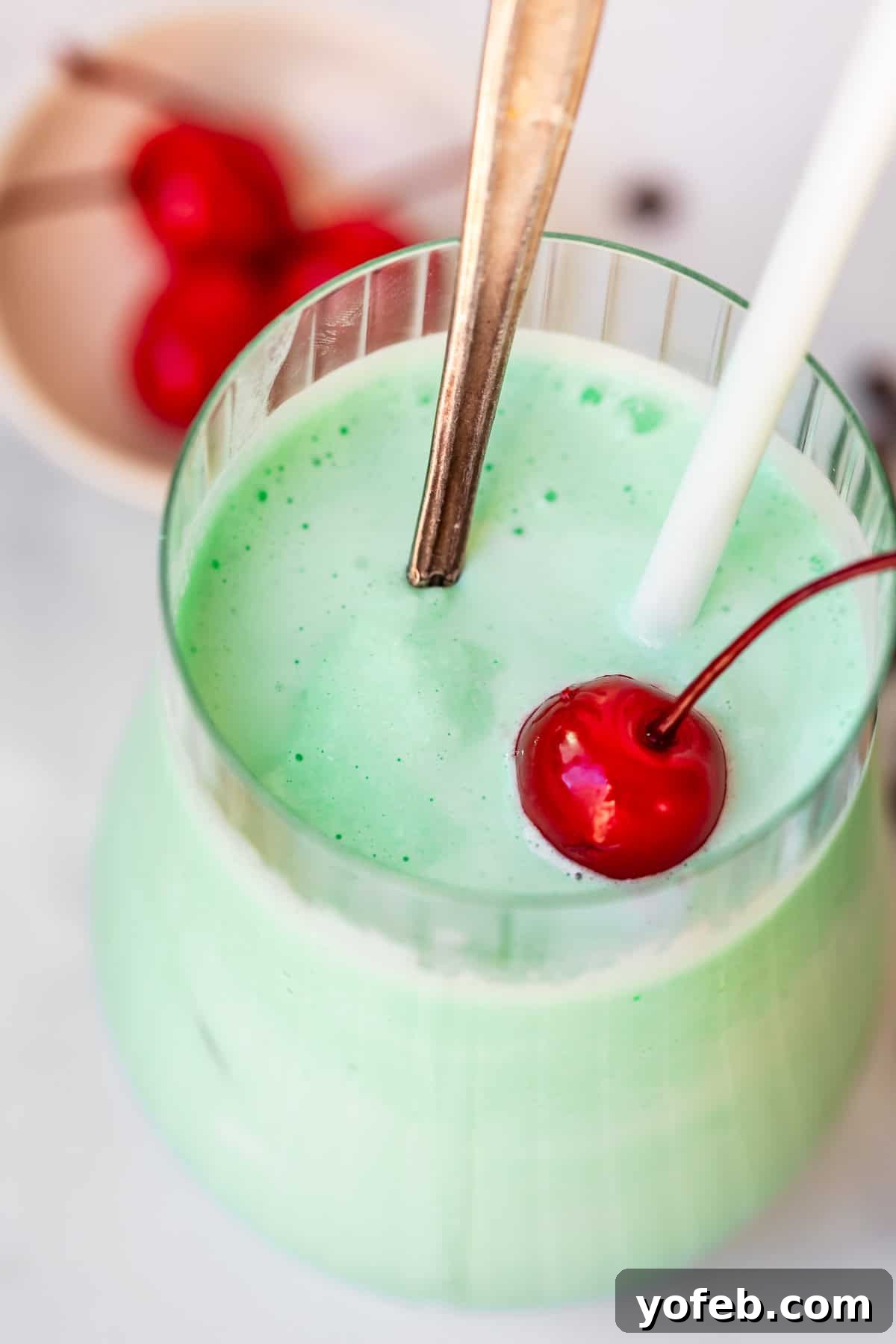 Leprechaun's Delight Shake 6 A close-up view looking into a glass cup with a perfectly blended Shamrock Shake, featuring a spoon, a straw, and a vibrant cherry. The texture looks incredibly smooth.