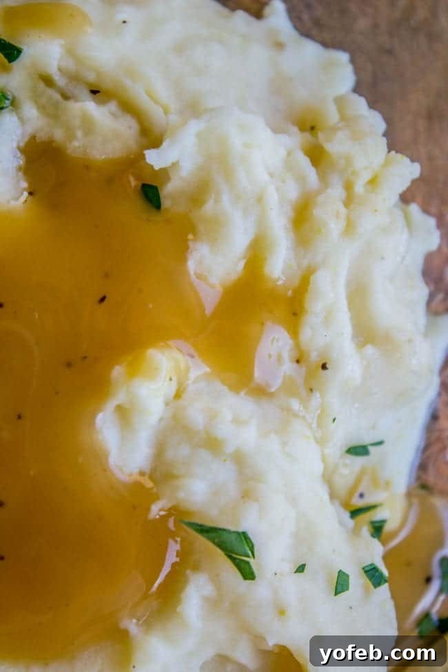 Creamy Mashed Potatoes: Aunt Shirley's Legendary Recipe - Fluffy, Creamy, Flavorful, Easy Homemade Side Dish for Thanksgiving, Pot Roast, or Any Meal. Made with Butter, Cream Cheese, and Garlic. A generous serving of fluffy mashed potatoes on a plate, with savory gravy generously poured over it.