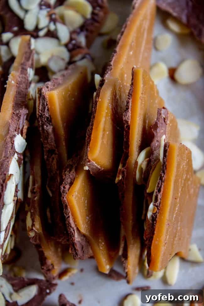 The Best Homemade English Toffee Recipe by The Food Charlatan. This is the BEST homemade English Toffee Recipe, hands down! This is such an easy and QUICK treat to make for the Christmas holidays. You don't even need a candy thermometer! #toffee #English #candy #Christmas #easy #homemade #tutorial #recipe #almonds Shards of English toffee stacked together on their side vertically, revealing layers of rich, caramelly candy, a smooth chocolate topping, and thinly sliced almonds, all presented on parchment paper.