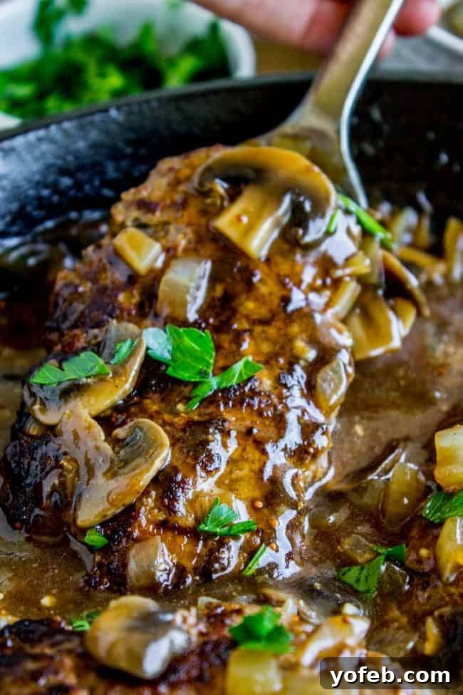 Best Salisbury Steak Recipe with Mushroom Gravy (30-Minute Dinner). Hearty ground beef patties pan-fried and served with a rich, flavorful mushroom gravy, fresh from the skillet. #ComfortFood #QuickDinner #SkilletRecipe #BeefDinner #MushroomGravy Close-up shot of Salisbury steaks swimming in rich mushroom gravy, garnished with vibrant fresh parsley, all served in a gleaming cast iron skillet. The texture of the gravy and the freshness of the herbs are prominent.