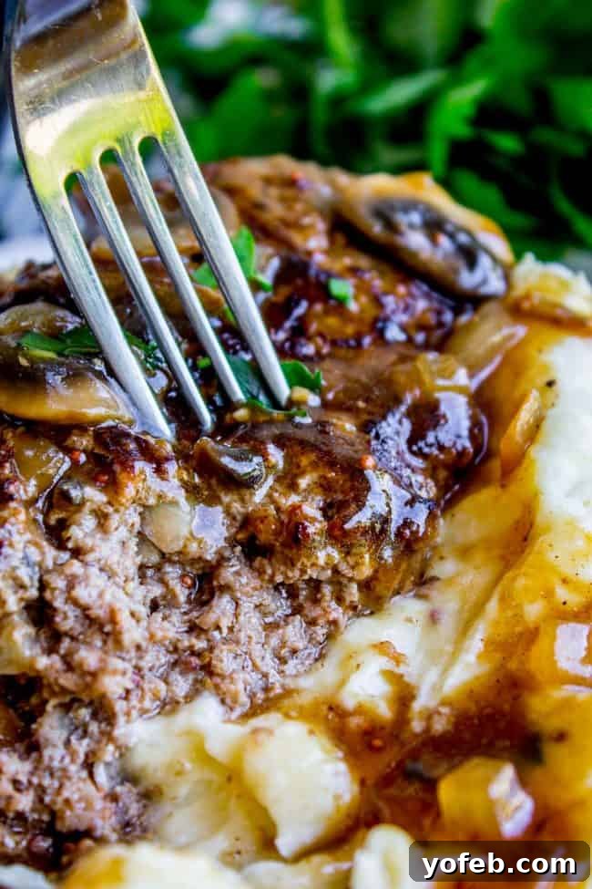 Salisbury Steak with Mushroom Gravy & Creamy Mashed Potatoes - A Comfort Food Classic. Enjoy the perfect bite of tender steak, rich gravy, and fluffy potatoes. #ComfortFoodGoals #SalisburySteakDinner #MushroomGravyRecipe #EasyMeal A fork lifts a generous bite of Salisbury steak, mushroom gravy, and creamy mashed potatoes from a plate, showcasing the textures and the irresistible combination of flavors.