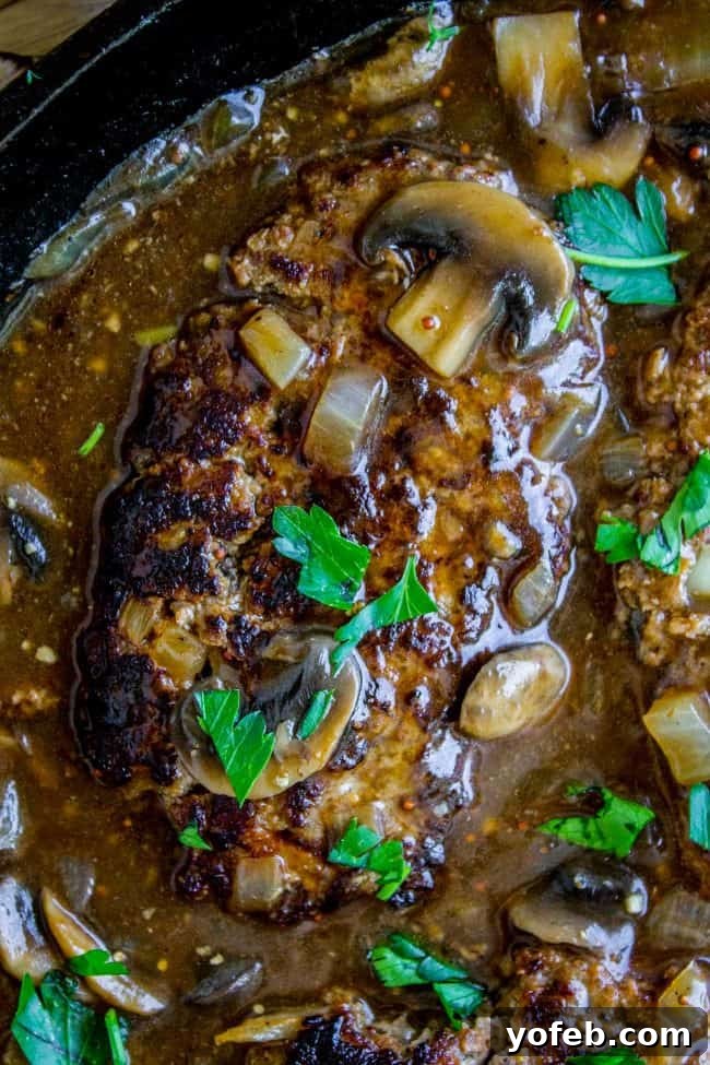 Skillet Salisbury Steak with Mushroom Gravy - Easy & Delicious 30-Minute Recipe. Hearty ground beef patties smothered in a rich, savory mushroom gravy, perfect for any occasion. #ComfortFoodClassics #EasyRecipe #GroundBeefDinner #MushroomGravy A close-up view of three Salisbury steaks richly coated in mushroom gravy, served warm in a cast iron skillet. The inviting texture of the gravy and the juicy appearance of the steaks are highlighted, ready to be enjoyed.