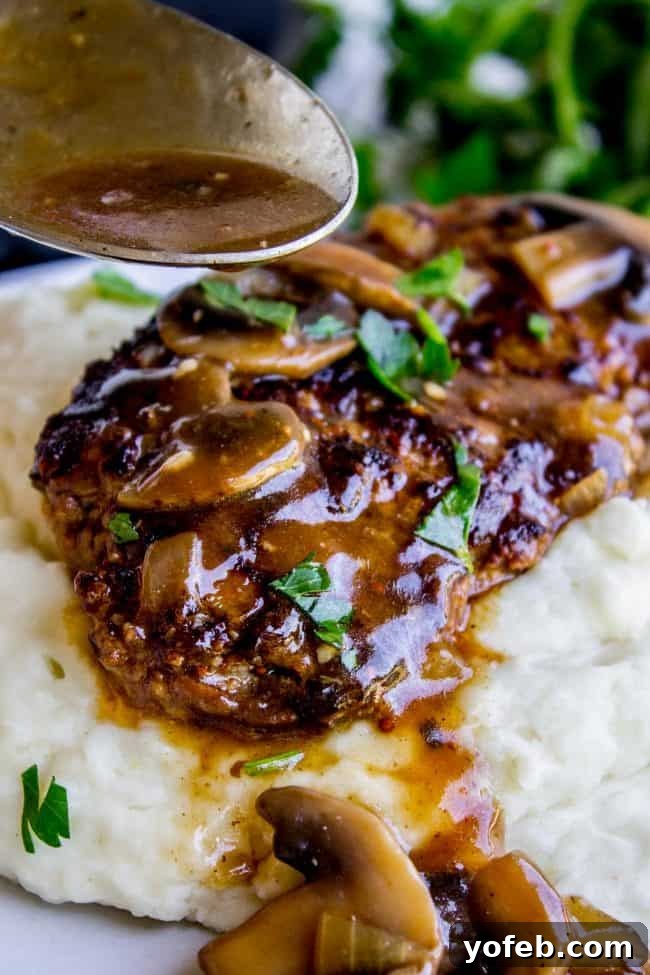 A juicy Salisbury Steak with sautéed mushrooms and brown gravy is served over creamy mashed potatoes, garnished with chopped parsley. A spoon drizzles extra savory gravy over the dish.