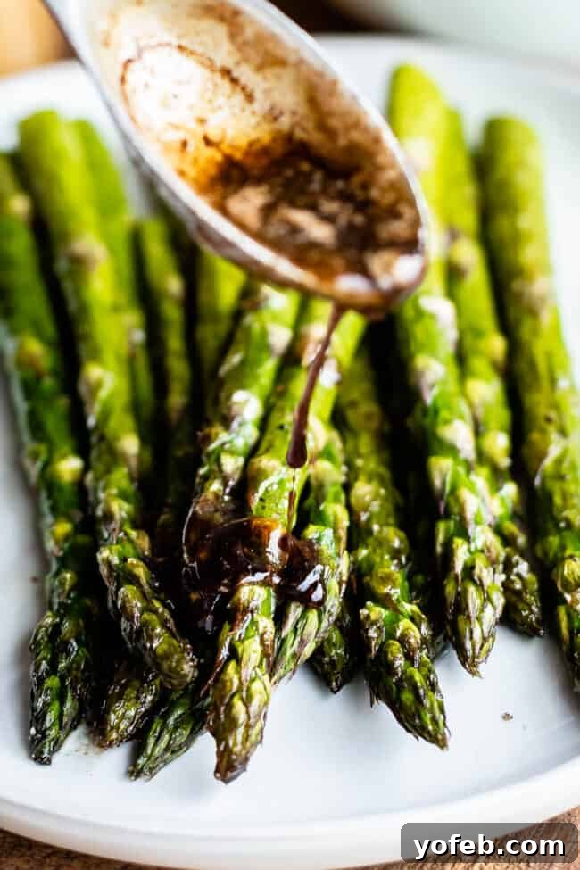 Brown Butter Balsamic Roasted Asparagus 3 Close-up of oven roasted asparagus spears on a white plate, drizzled with a dark balsamic sauce from a spoon.