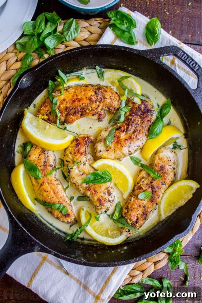 A black skillet filled with golden-brown lemon basil chicken in a creamy sauce, garnished with fresh basil and lemon wedges, sits on a checkered towel on a wooden table.