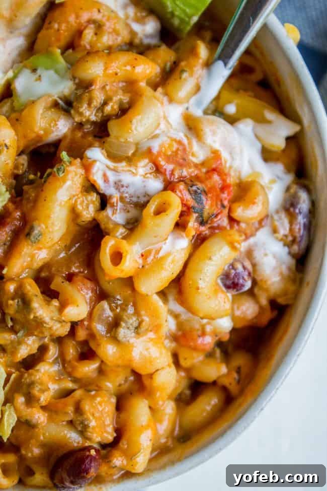 Creamy Chili Mac and Cheese with Sour Cream A close-up shot of a bowl of creamy Chili Mac and Cheese, garnished with a dollop of sour cream, being scooped with a spoon.