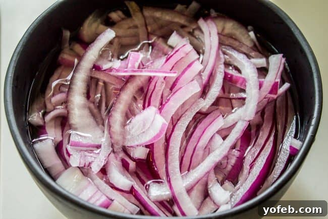 Prepping red onions for Greek salad: A simple trick to remove harshness and enhance flavor. #redonion #preptips #greeksalad #easyrecipe #cookinghacks Thinly sliced red onions soaking in a bowl of cold water to reduce their pungent bite, a preparatory step for a fresh Greek salad.