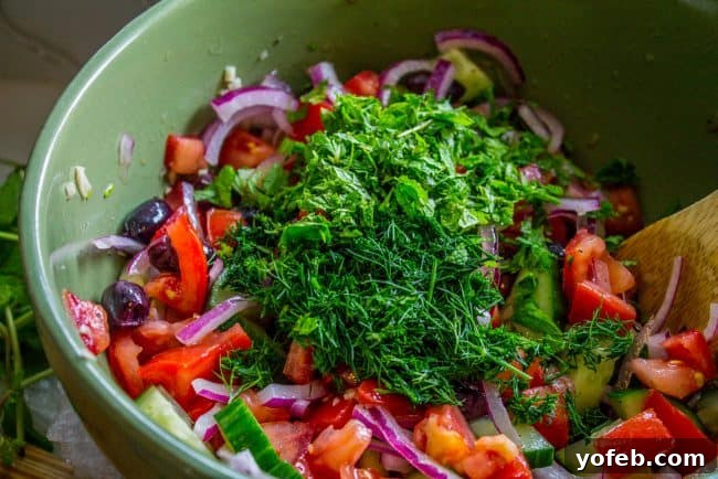 Adding fresh herbs to Greek salad: Chopped dill and mint bring unparalleled freshness and aromatic depth to this healthy and easy recipe. #freshherbs #dill #mint #greeksalad #flavorboost Freshly chopped dill and mint are mixed with other vibrant ingredients in a green bowl, preparing a homemade Greek cucumber tomato salad with generous feta cheese.
