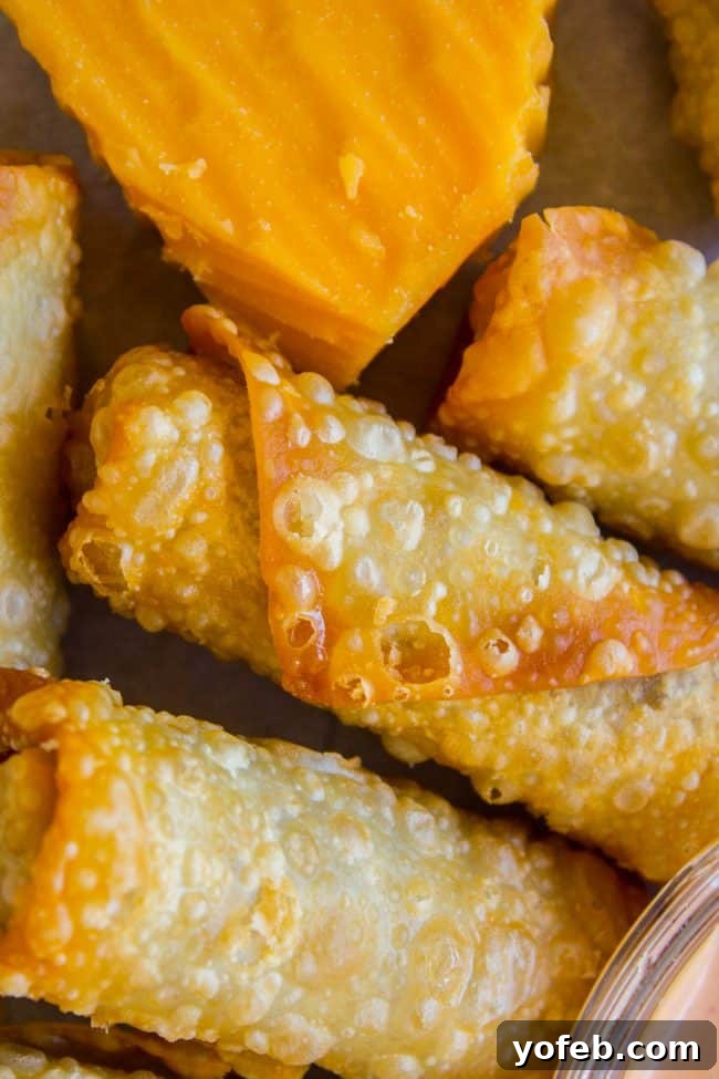 Cheeseburger Egg Rolls from The Food Charlatan. These have got to be one of the best mashups of all time! I mean, cheesy hamburger filling that's deep fried in wrappers?? How can we go wrong here? Dip it in a delicious fry sauce and you will be in cheeseburger heaven in no time! #cheeseburger #burger #hamburger #cheesy #cheese #fried #deepfried #easy #recipe #beef #homemade #howtomake #frysauce #appetizer #dip Crispy cheeseburger egg rolls arranged next to a block of fresh cheddar cheese.