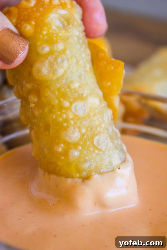 Cheeseburger Egg Rolls from The Food Charlatan. These have got to be one of the best mashups of all time! I mean, cheesy hamburger filling that's deep fried in wrappers?? How can we go wrong here? Dip it in a delicious fry sauce and you will be in cheeseburger heaven in no time! #cheeseburger #burger #hamburger #cheesy #cheese #fried #deepfried #easy #recipe #beef #homemade #howtomake #frysauce #appetizer #dip A hand dipping a golden-brown cheeseburger egg roll into a rich, creamy fry sauce.
