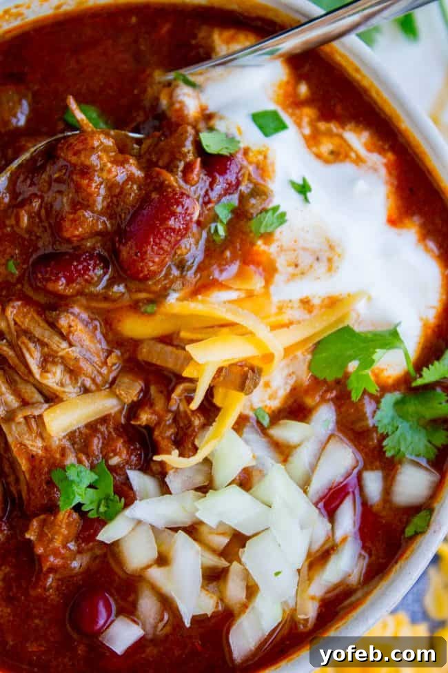 The Best Shredded Beef Slow Cooker Chili - A hearty, award-winning recipe featuring tender, fall-apart beef and a unique touch of Masa Harina for incredible flavor. A hearty bowl of slow cooker chili, generously topped with shredded cheddar cheese, a dollop of sour cream, finely chopped red onions, and fresh cilantro, photographed from an overhead perspective.