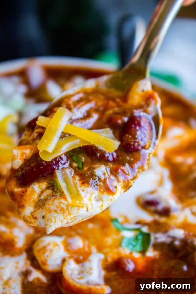 Spoonful of the Best Chili Ever - Highlighting the rich texture and delicious toppings. A close-up shot of a spoonful of slow cooker chili being lifted from a bowl, showcasing the tender shredded beef, beans, and rich sauce, topped with melted cheddar cheese, sour cream, and fresh cilantro.