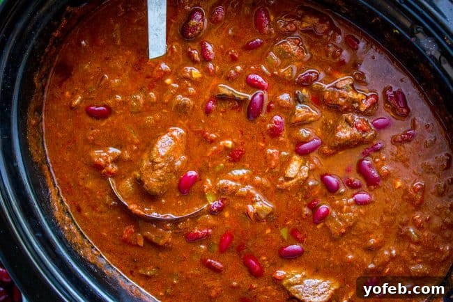 Chili simmering in a crockpot, prepared for serving. Overhead shot of rich beef chili in a crock pot, stirred with a metal ladle, ready to be served.