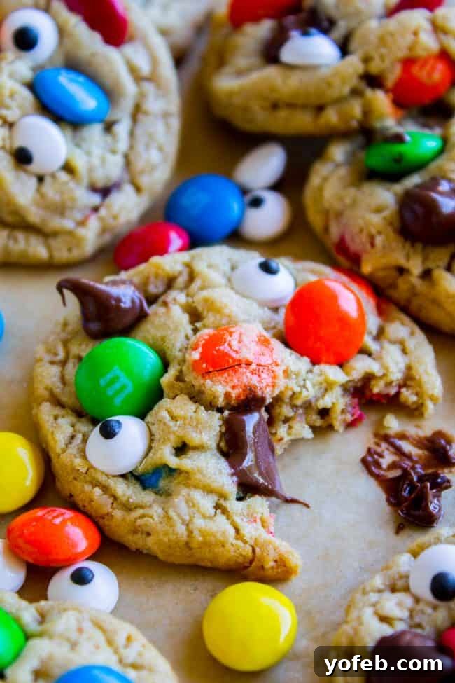 Close-up of monster cookies topped with candy-coated chocolates and candy eyes, one cookie partially eaten, with melted chocolate chips and scattered candies on a light surface.