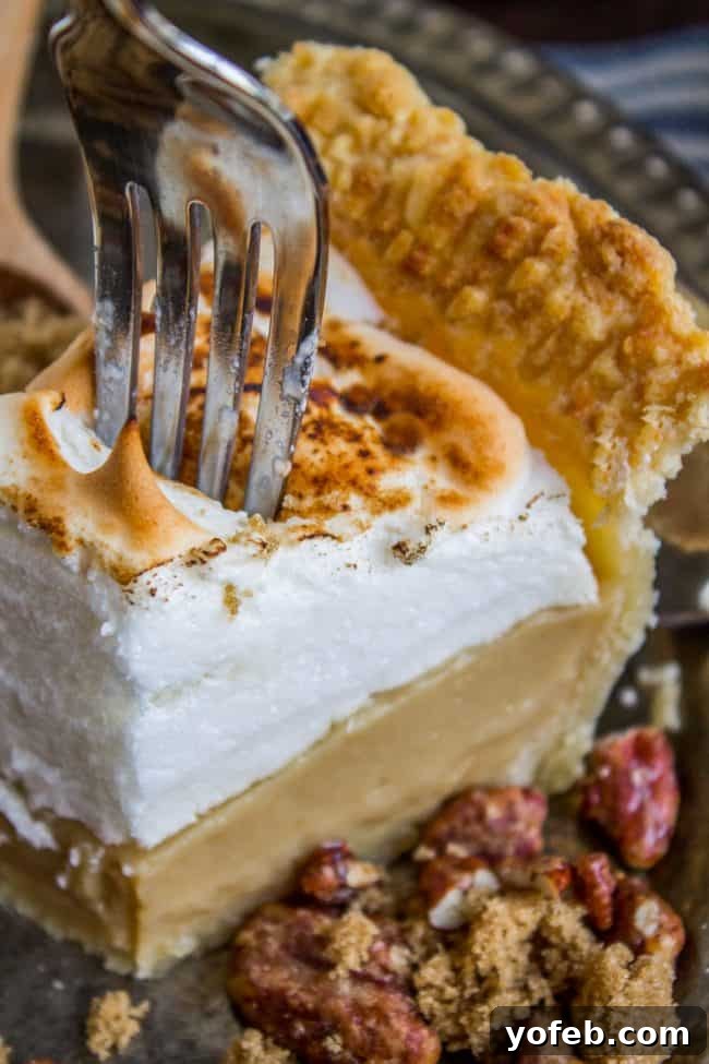 A fork cuts into a slice of butterscotch pie with a golden-brown crust, fluffy toasted meringue, and creamy filling. Crumbled pecans and brown sugar are scattered on the plate beside the pie.
