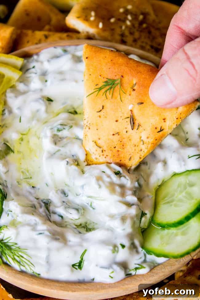 Tzatziki Sauce Recipe (with Toasted Za'atar Pita: Easy Appetizer!) from The Food Charlatan. Have you ever tried making a Greek Tzatziki sauce recipe? Creamy Greek yogurt is combined with cool cucumbers and spiced with garlic, dill, and lemon. It is simple, fresh, and healthy. I paired it with toasted pita bread to make an easy New Year's Eve appetizer! #Greek #tzatziki #sauce #cucumber #lemon #dill #garlic #recipe #uses #appetizer #oliveoil #dip #healthy #gyro #easy #rezept #greekyogurt #lunch A hand dipping a piece of pita bread into creamy cucumber tzatziki sauce, garnished with fresh dill.