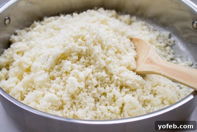 Cauliflower rice being cooked in a large pan with a wooden spoon, seasoned to perfection.