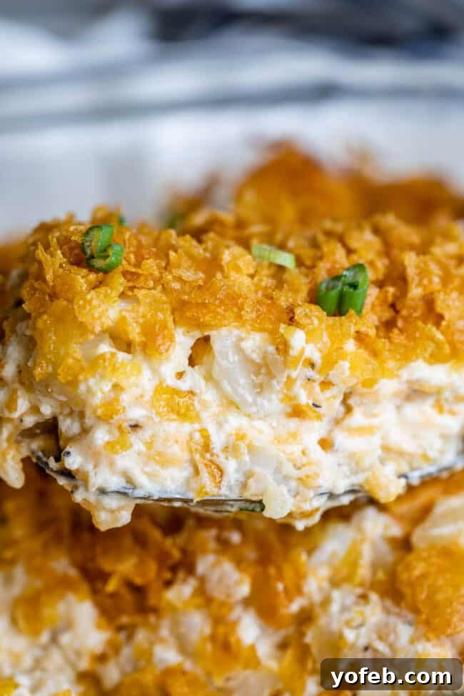 Funeral Potatoes (Cheesy Potato Casserole) from The Food Charlatan. If you haven't heard of this side dish, you will love it! Potatoes mixed with sour cream, butter, white sauce (or a can of soup - I include instructions for both), and tons of cheddar cheese, then baked to perfection with a crunchy corn flake topping. It doesn't get any better for holidays! #potatoes #withhashbrowns #easy #casserole #baked #cornflakes #fromscratch #withcornflakes #inoven #soup #homemade #quick #best spoon lifting cheesy potato casserole from dish, topped with green onions.