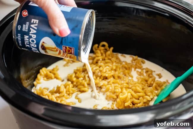 A can of evaporated milk being poured into a slow cooker containing dry noodles and other liquid ingredients.