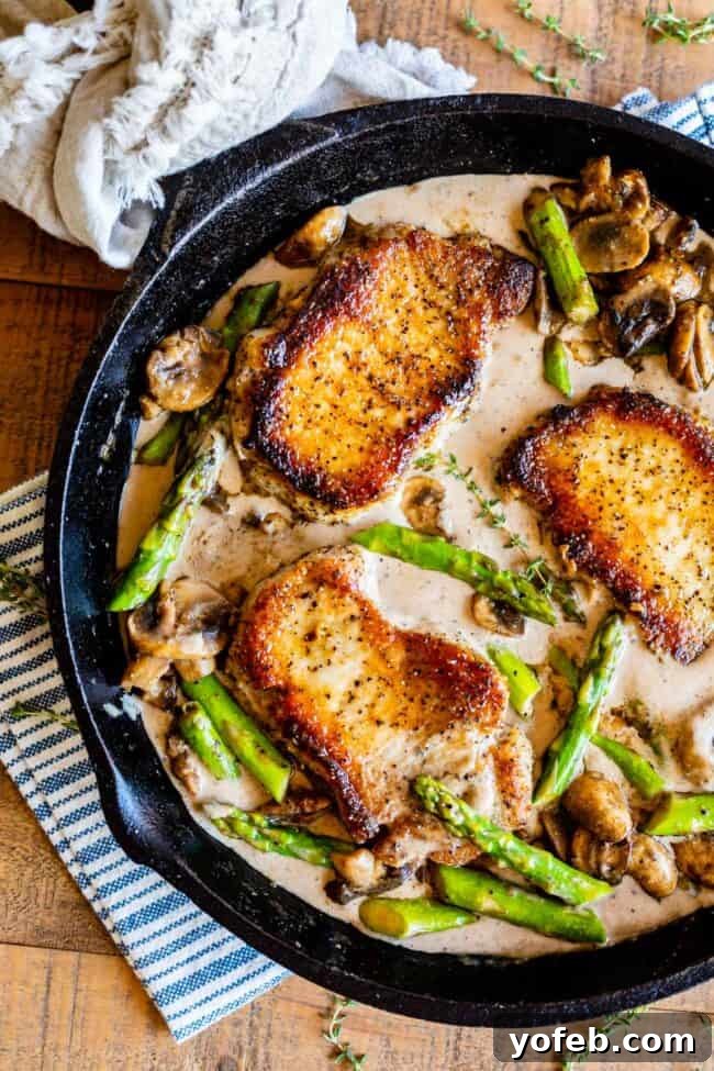 Pan-Seared Pork Chops with Velvety Mushroom Sauce 2 Juicy boneless pork chops with a golden-brown crust, served alongside vibrant green asparagus and tender sautéed mushrooms, all generously coated in a creamy, savory pan sauce, presented in a cast iron skillet.
