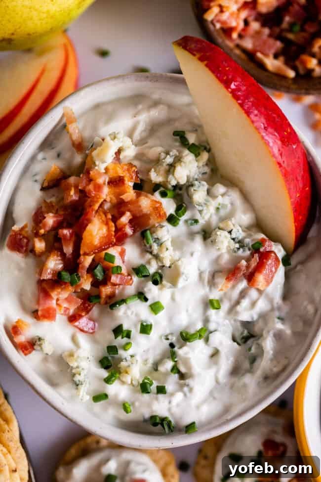 Ultimate Bacon Blue Cheese Dip 3 A bowl of creamy blue cheese dip topped with crumbled bacon and chopped chives, garnished with a sliced red apple. Crackers and apple slices surround the bowl.