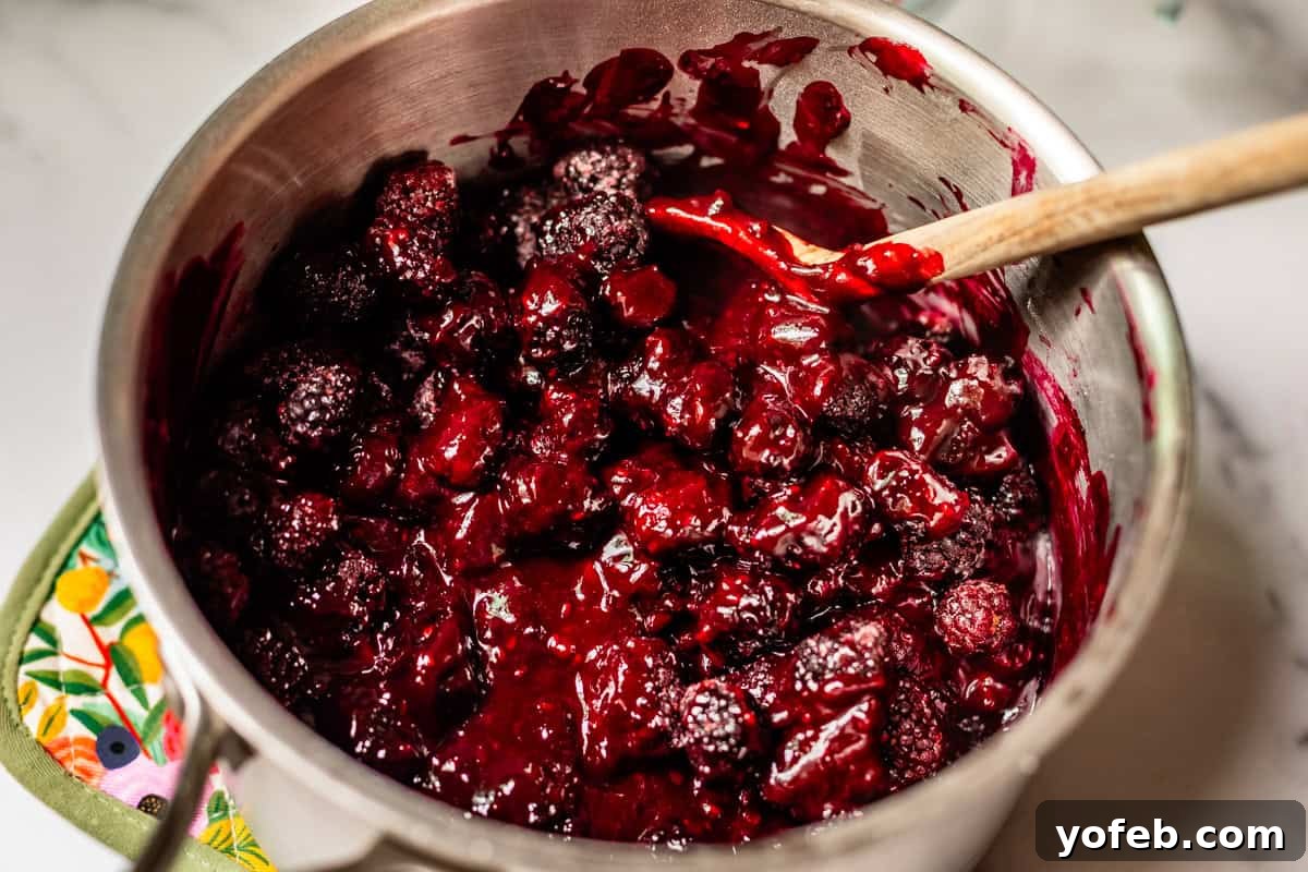 Grandma Georgias Farmhouse Blackberry Bake 15 A stainless steel saucepan filled with thick, glossy mixed berry compote, with whole blackberries visible, being stirred by a wooden spoon on a white countertop.