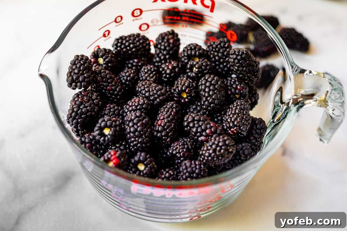 Grandma Georgias Farmhouse Blackberry Bake 16 A glass measuring cup filled with fresh blackberries sits on a white surface, with a few blackberries scattered in the background.