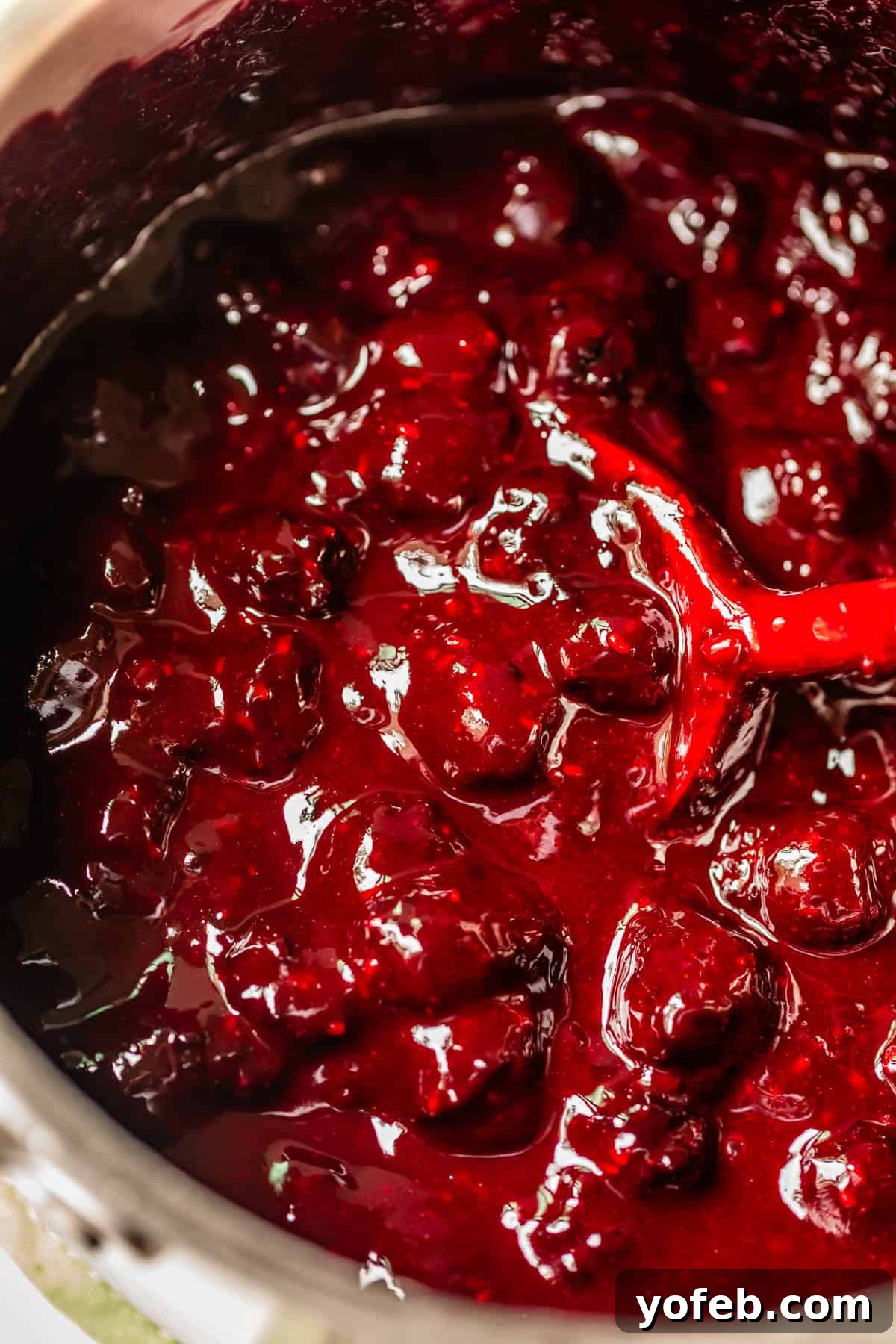 Grandma Georgias Farmhouse Blackberry Bake 18 Close-up of a pot filled with thick, glossy blackberry pie filling, with whole berries and juices visible. A red spoon is partially submerged, stirring the mixture.