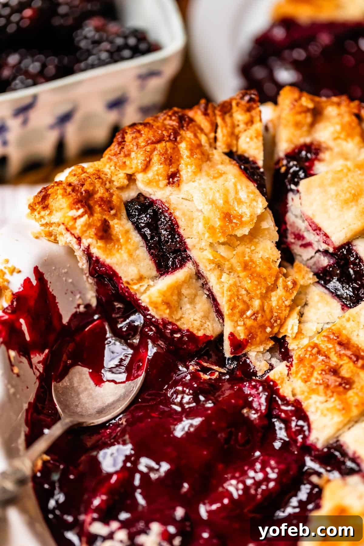 Grandma Georgias Farmhouse Blackberry Bake 24 A close-up of a homemade blackberry pie with a golden, flaky lattice crust. A slice is being served, revealing juicy blackberry filling and a spoon resting in the pie dish. Fresh berries are seen in the background.