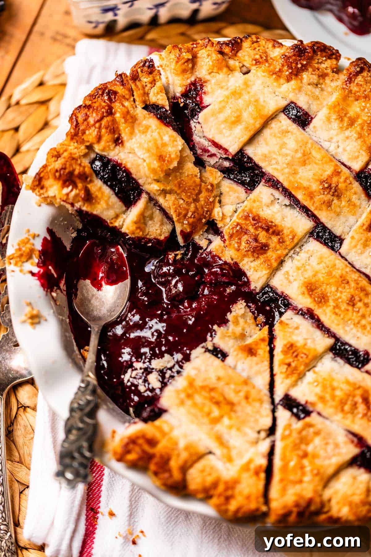 Grandma Georgias Farmhouse Blackberry Bake 5 A close-up of a homemade blackberry pie with a golden lattice crust, partially sliced. A silver spoon rests in the pie dish, revealing the juicy berry filling. The pie sits on a white cloth with a woven placemat underneath.