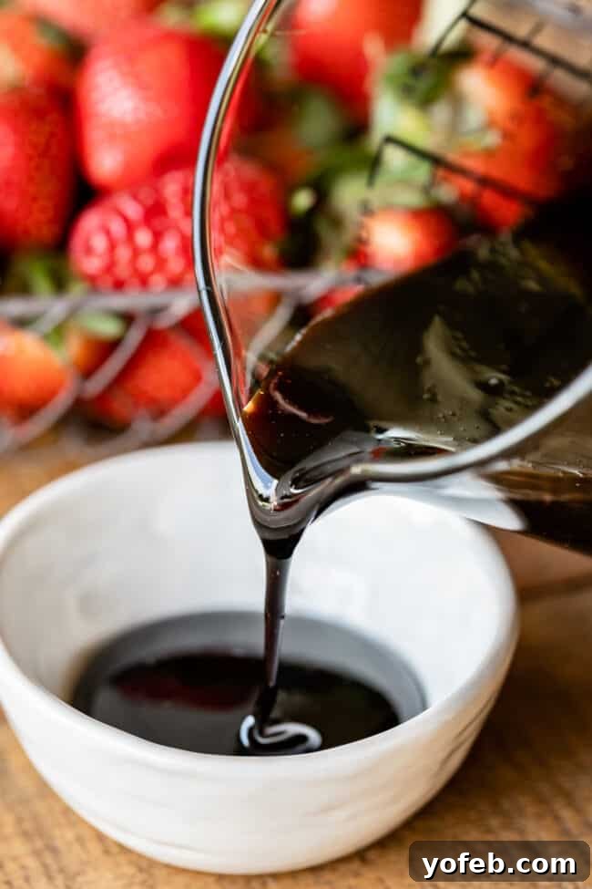 Sweetened Balsamic Glaze for Desserts Pouring homemade balsamic vinegar reduction from a glass measuring cup into a small white ceramic bowl, with fresh strawberries in the background.