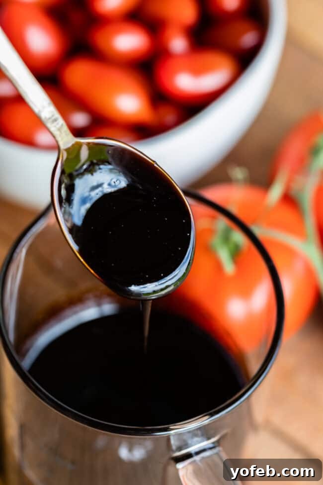 How to Identify Perfect Balsamic Glaze Consistency Rich, syrupy balsamic reduction drizzling from a metal spoon into a glass measuring cup, with fresh tomatoes in the background.